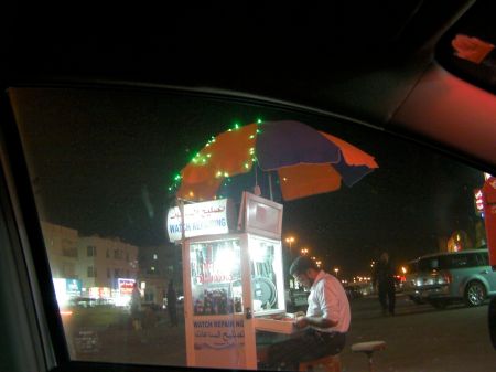 Watch Repair Kiosk man sitting at a small booth under a large umbrella doing watch repair