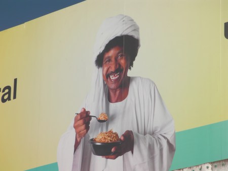 close up of man pictured on the billboard in Middle Eastern clothing and turbin, grinning big as he eats food