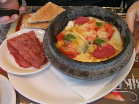 clay pot containing layered bread, fried eggs, tomatoes, bell peppers, pepperoni, cheese