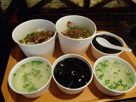 bowls of rice noodles with toppings, bowls of broth, and bowls of black gelatin