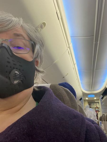 woman wearing black medical mask on a plane, at the back of the plane are medical inspectors wearing hazmat suits