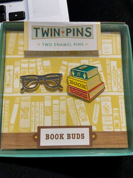 a box containing two pins, one shaped like eyeglasses, the second shaped like a pile of three books