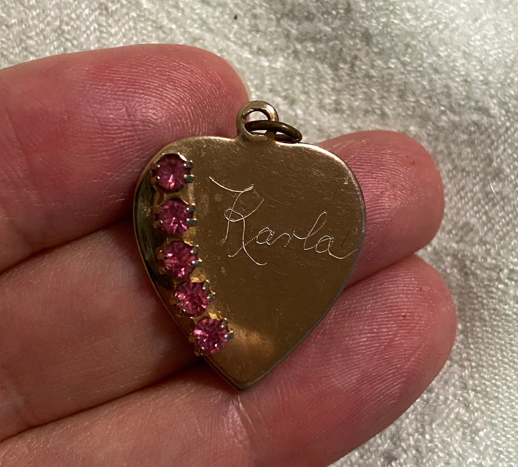fingers holding a heart shaped pendant with five pink stones and a name inscribed