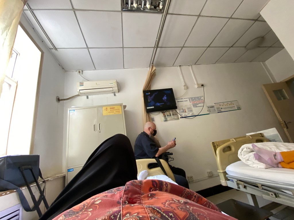 small hospital room with two occupied beds, man sitting on a wheelchair