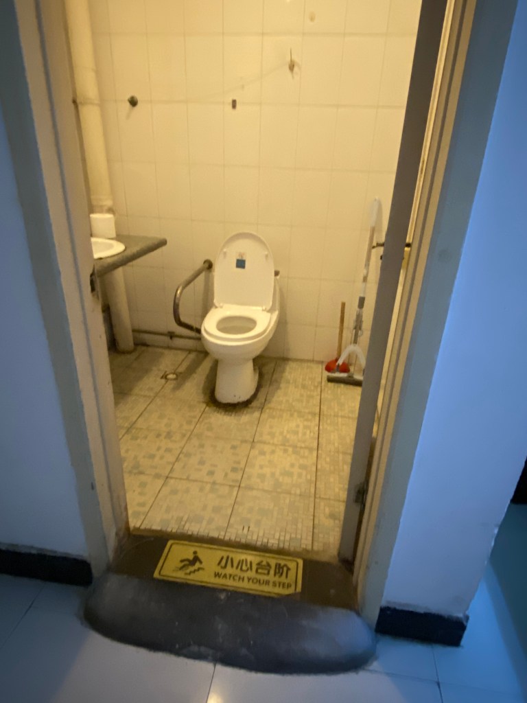 toilet and sink, warn floor tile, brown stuff around the base of the toilet, a plunger and mop in the corner, a cement ramp at the entrance